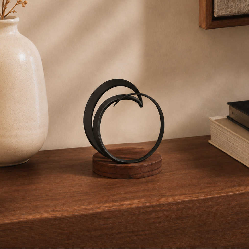 A black metal abstract sculpture on a round wooden base, styled on a wooden shelf next to a cream ceramic vase and books against a beige wall.