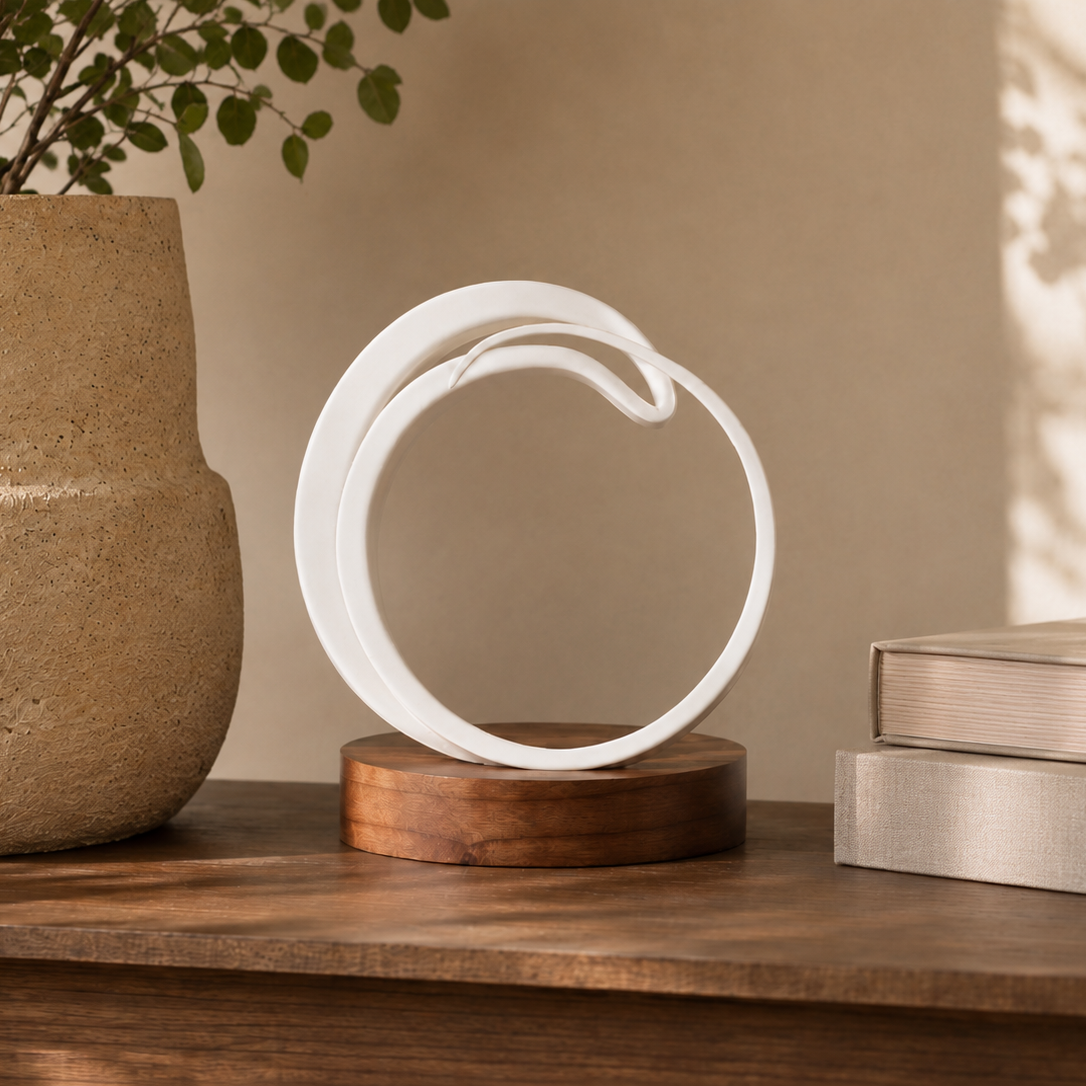 A white sculptural ring-shaped decor piece displayed on a round wooden base, set on a wooden shelf next to a large textured beige planter and stacked books.